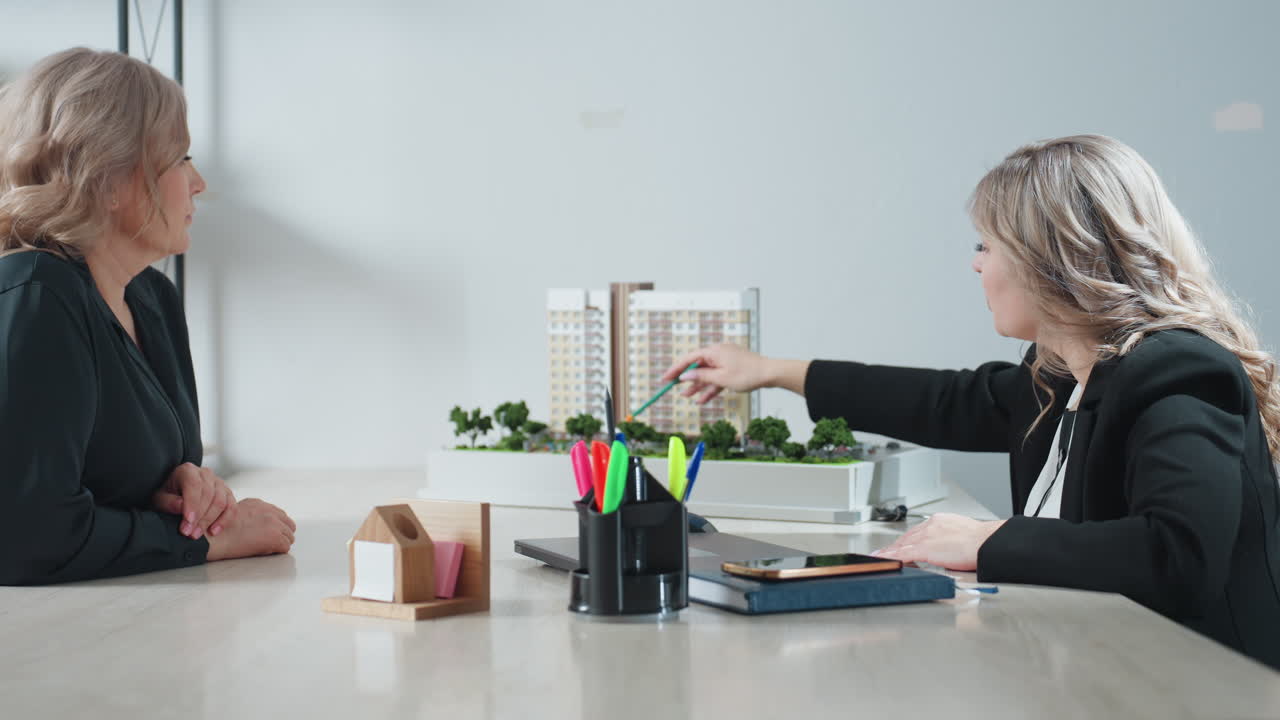 saleswoman gestures with pencil toward architectural building miniature while explaining property features to unseen client during real estate meeting, emphasizing structure details and layout