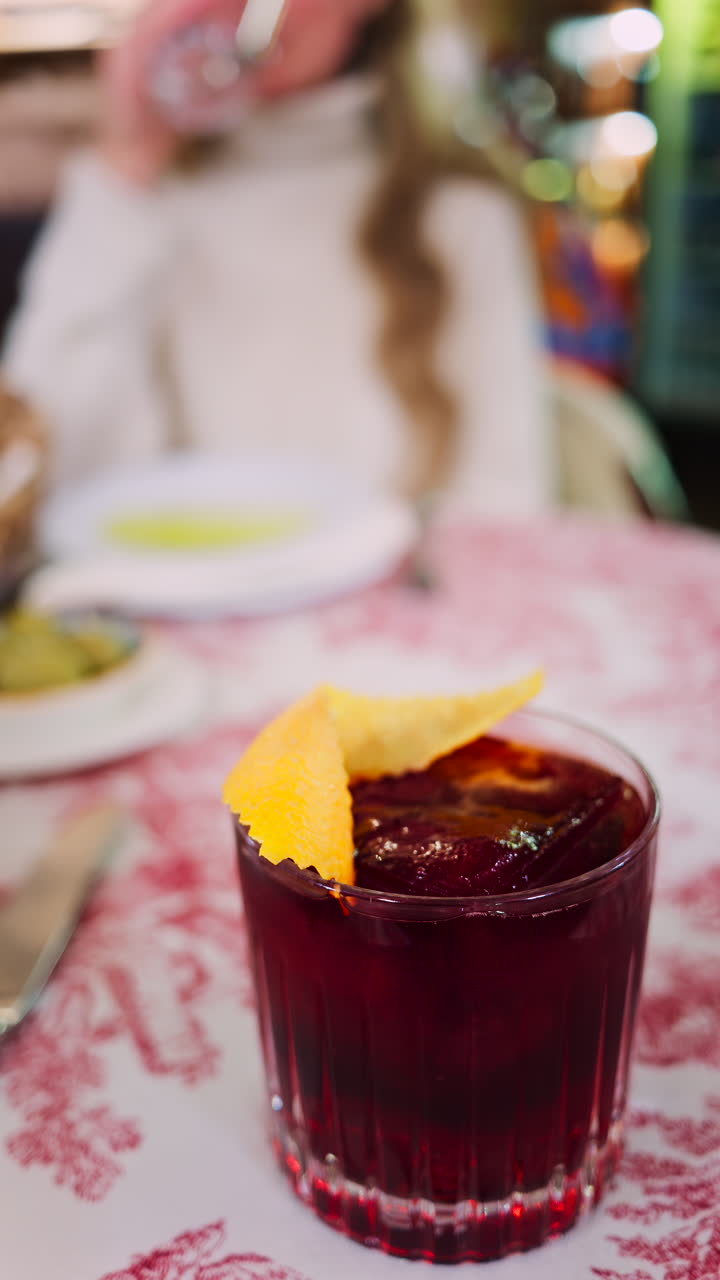 Close up of a negroni cocktail on a red and white tablecloth with a woman drinking at a restaurant on the background. Vertical