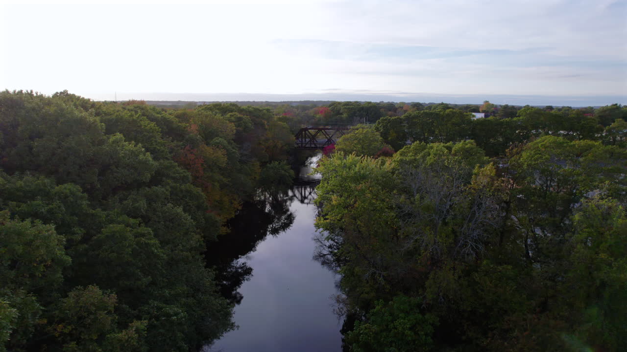 Aerial drone video of the peaceful Pawtuxet River in Cranston, Rhode Island lined with color changing fall foliage and bookended by a railroad bridge and a road bridge