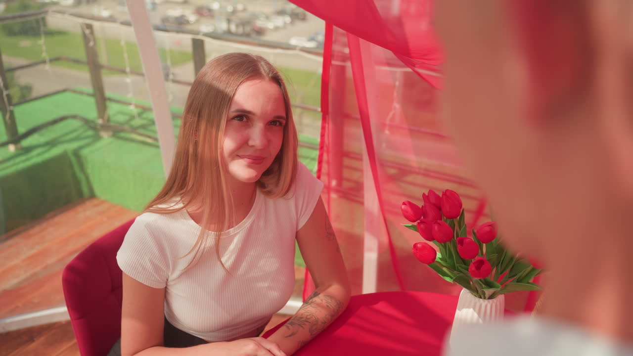 Giovane coppia bianca ad un appuntamento sul balcone, donna seduta a un tavolo rosso che sorride al partner, vaso di tulipani rossi, tende leggere e vista sulla città illuminata dal sole, atmosfera calda e accogliente con conversazione giocosa e sguardo tenero