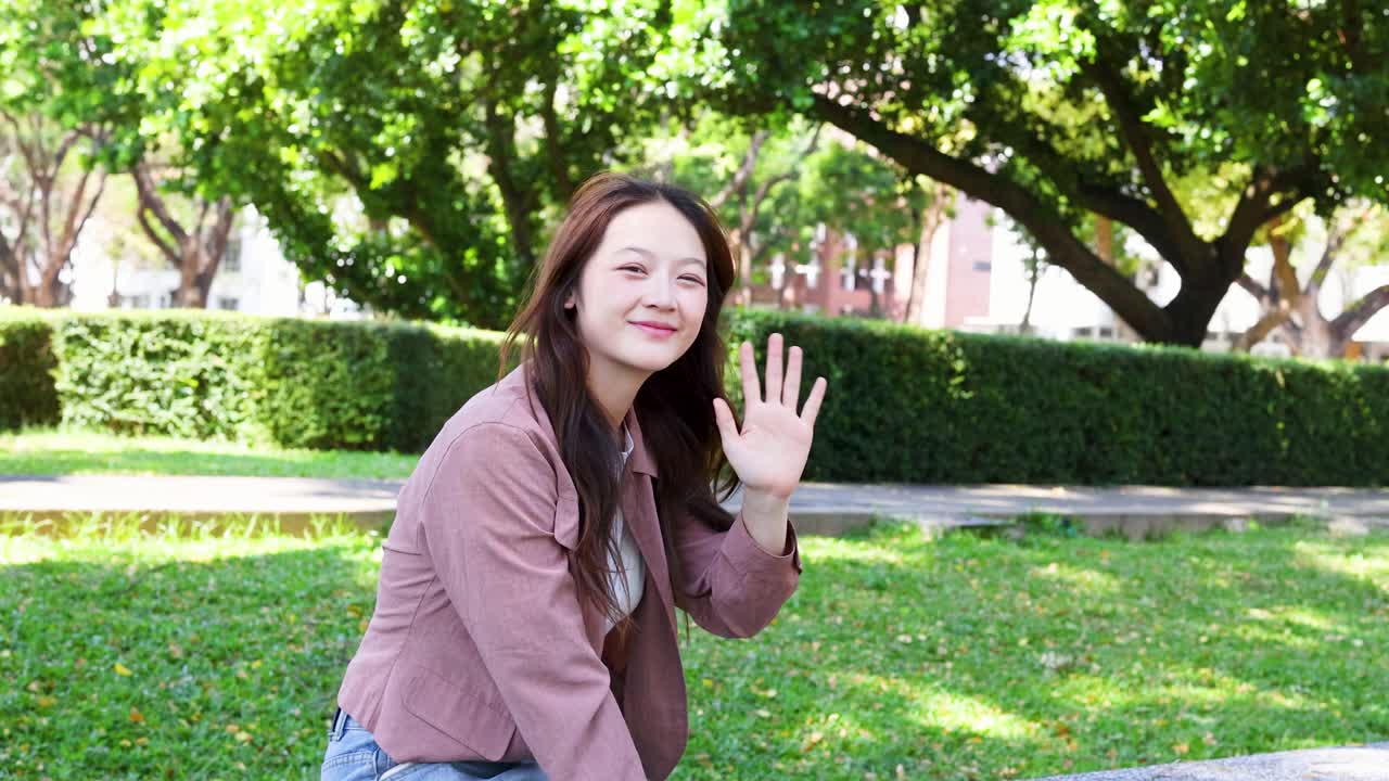 분홍색 블레이저를 입고 캠퍼스 잔디밭에서 카메라를 향해 손을 흔드는 미소 짓는 태국 학생
