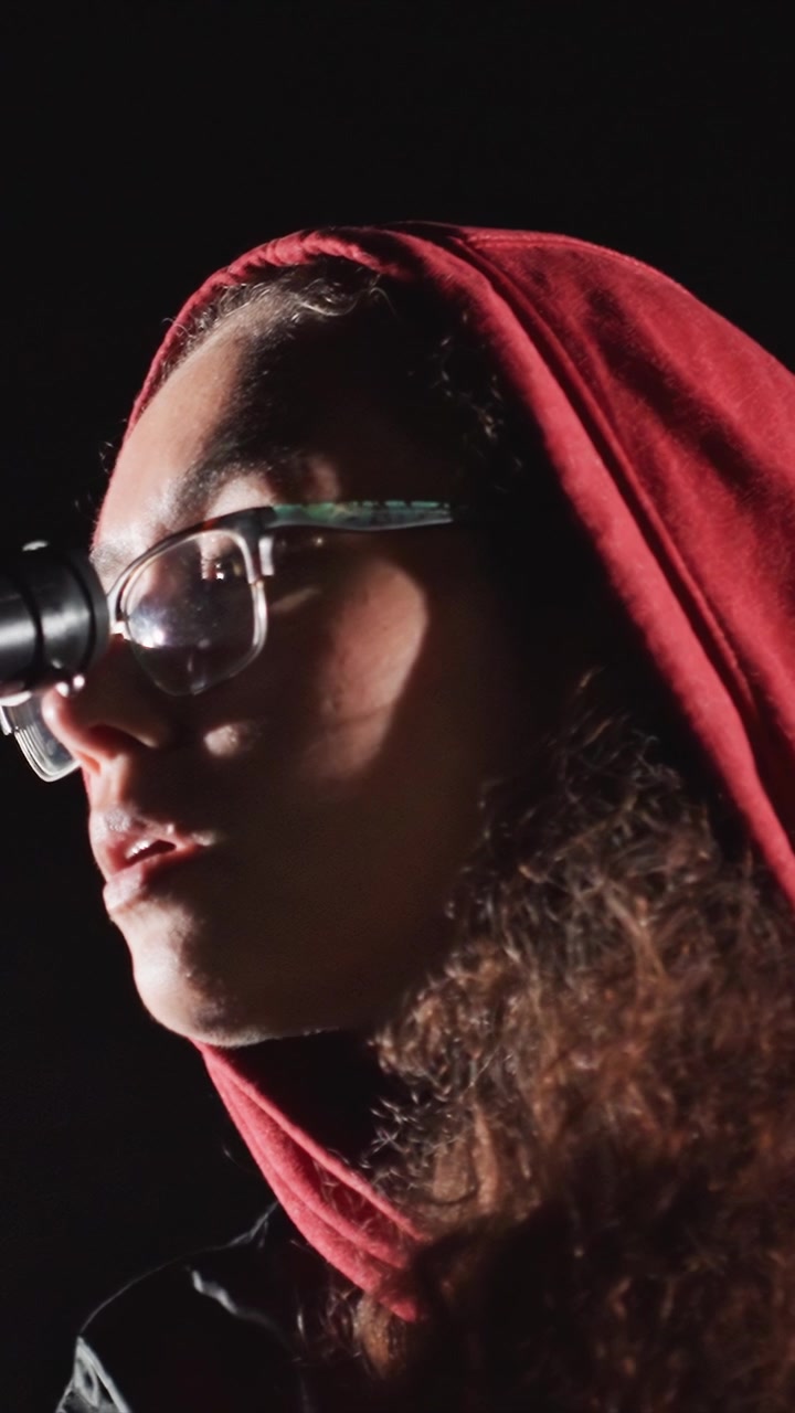 Curly haired woman speaks into microphone wearing. Black framed eyewear rests upon face of person wearing. Black cylindrical microphone visible on stand near speaker left