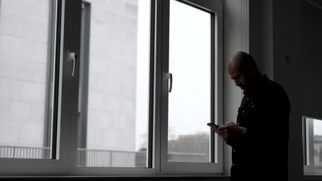 Un-Hombre-Está-Parado-Junto-A-La-Ventana-Con-Un-Clima-Sombrío-Afuera,-Absorto-Escribiendo-En-La-Pantalla-De-Su-Teléfono-Móvil