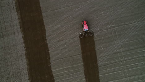 Red-tractor-with-harrows-during-plowing-brown-field-with-green-grass-around