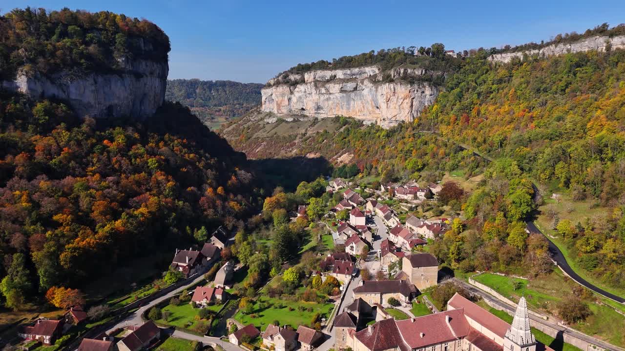 Aerial view of Baume les Messieurs, France, with its village positioned between tall limestone cliffs and dense autumn forests, winding roads, rustic rooftops, and scenic countryside