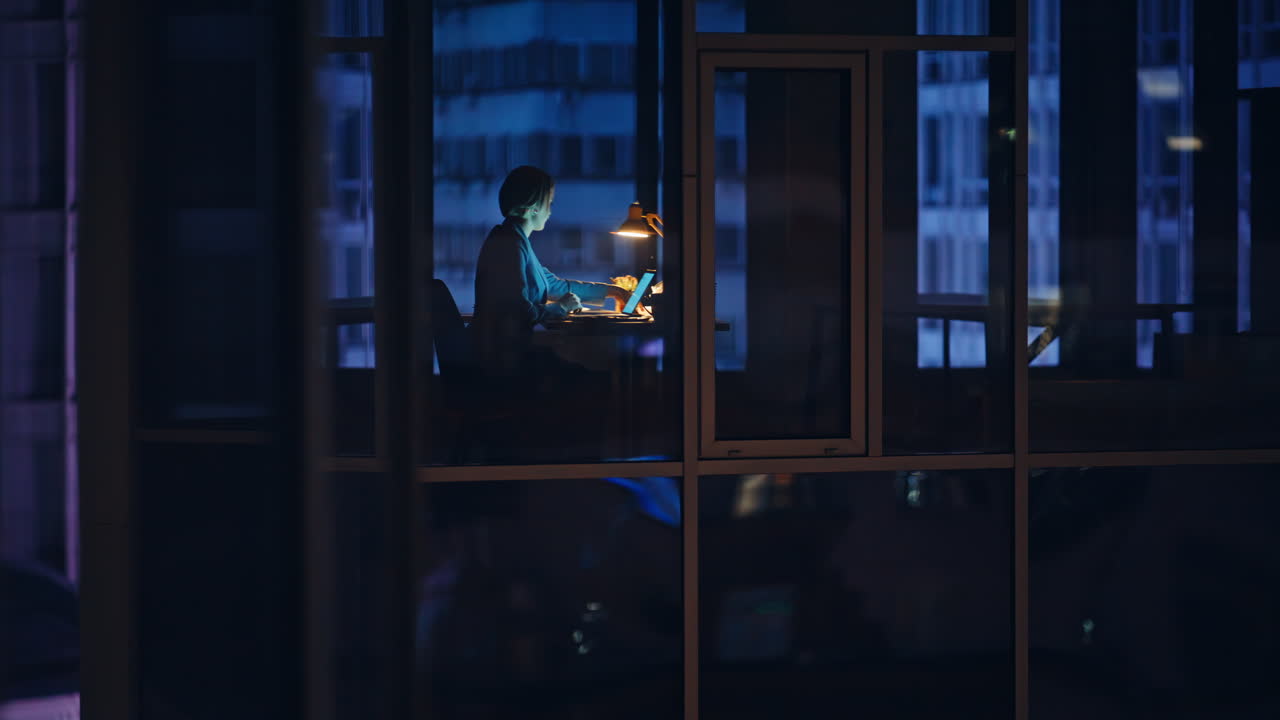 Glass wall view employee browsing laptop during night shift. Office specialist