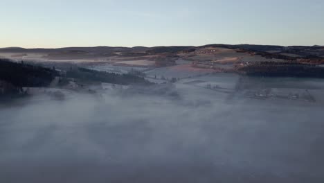 Vista-Aérea-Del-Sereno-Paisaje-De-Colinas-Y-Valles,-Parcialmente-Envuelto-En-Niebla-Bajo-La-Suave-Luz-Del-Amanecer.