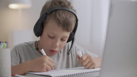 Un-Niño-Pequeño-Está-Charlando-En-Línea-Con-Auriculares-Con-Micrófono-Mirando-La-Pantalla-De-La-Computadora-Portátil-Y-Dibujando-Sentado-En-La-Mesa-En-Casa