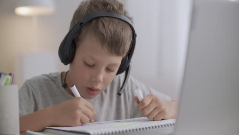 little boy is chatting online by headphones with microphone looking at screen of laptop and drawing sitting at table at home