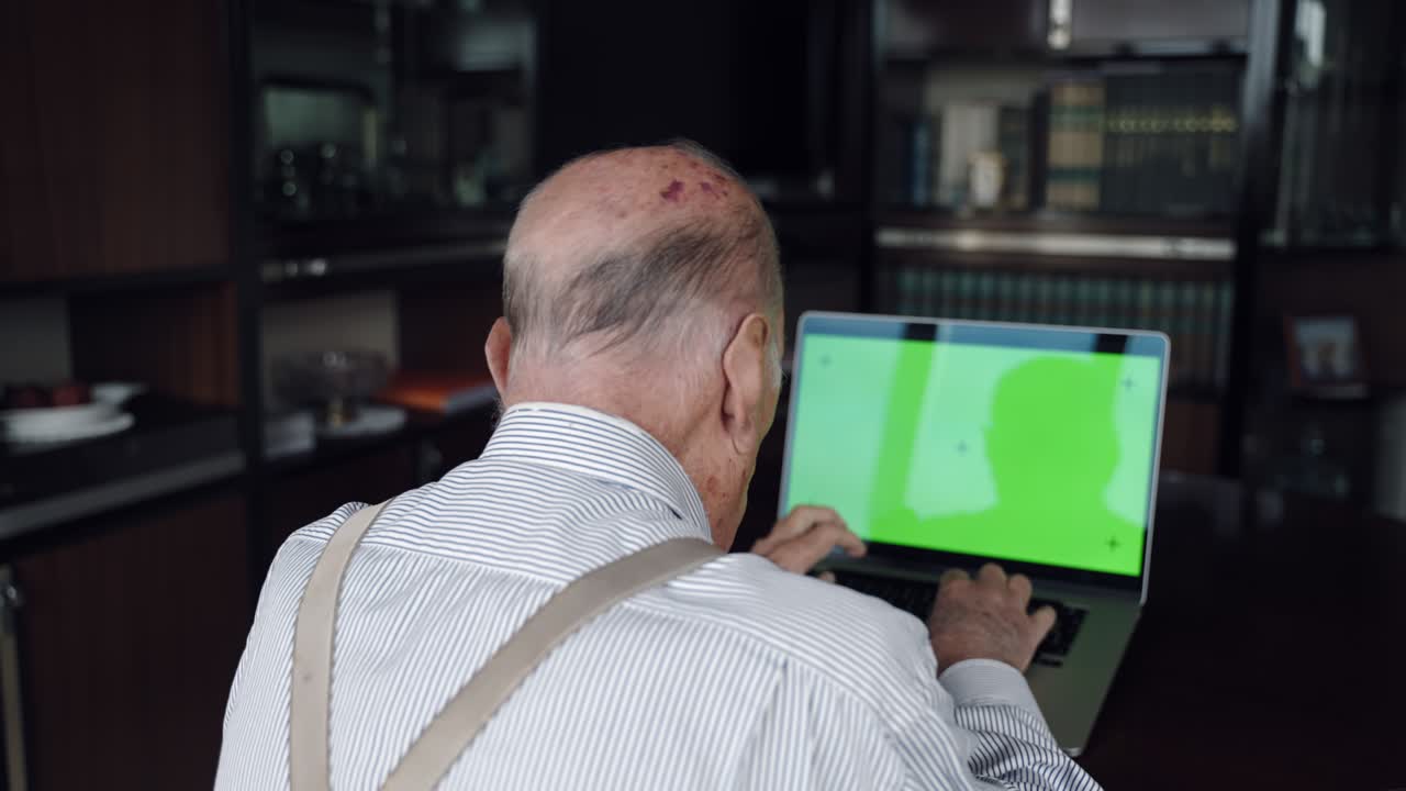 vista trasera de un anciano jubilado trabajando en casa con una computadora portátil de pantalla verde, un anciano sentado en la mesa y escribiendo en el teclado de la computadora.