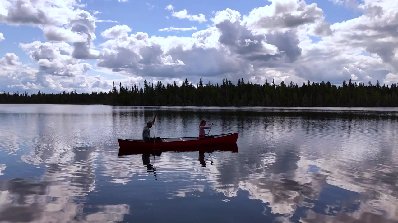 Ein Paar beginnt eine Kanufahrt über einen ruhigen See, der Wolken unter einem hellen Himmel widerspiegelt, umgeben von friedlichem Wald in der Wildnis Nord-Ontarios.