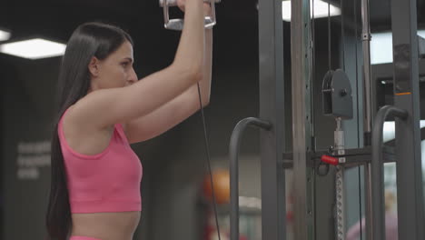 a hispanic brunette woman in a pink suit pulls a rope in a crossover with her hands to train her shoulders. shoulder workout in a trainer. professional woman instructor