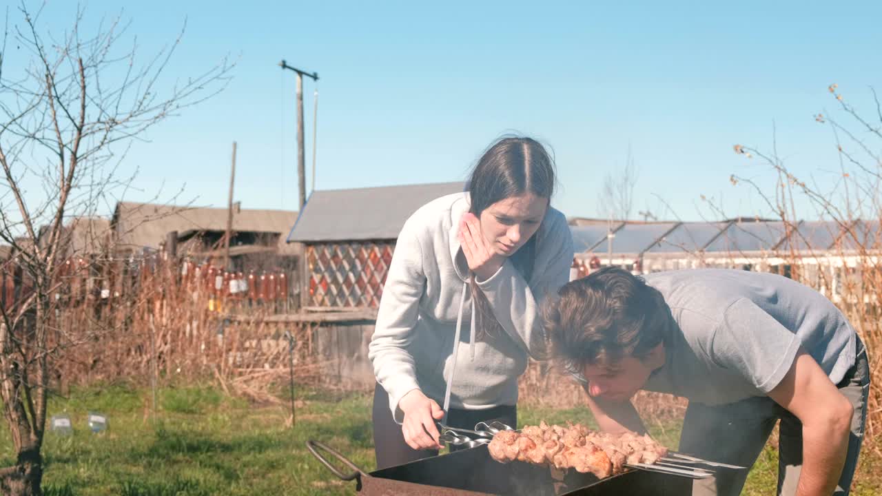 남자와 여자가 뒷마당의 목탄 그릴 위에 샤슬릭 고기를 요리합니다. 함께 얘기하고 미소 짓고 있습니다.