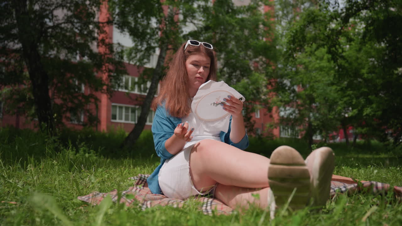 Young woman sitting on grass during bright summer day, focused on embroidery hoop while stitching floral pattern, enjoying peaceful outdoor atmosphere filled with sunlight