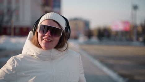 close up of performer smiling brightly in white winter coat and sunglasses, expressing joy through motion outdoors, with sunlight highlighting her face as she moves in cold city environment
