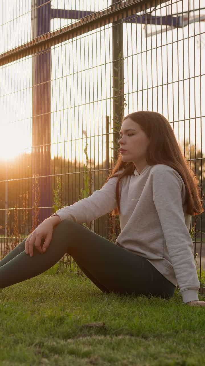 una joven se sienta en la hierba en la puesta de sol desvanecida, luchando con sentimientos de soledad y frustración durante un momento desafiante y desalentador de la vida de la mujer.