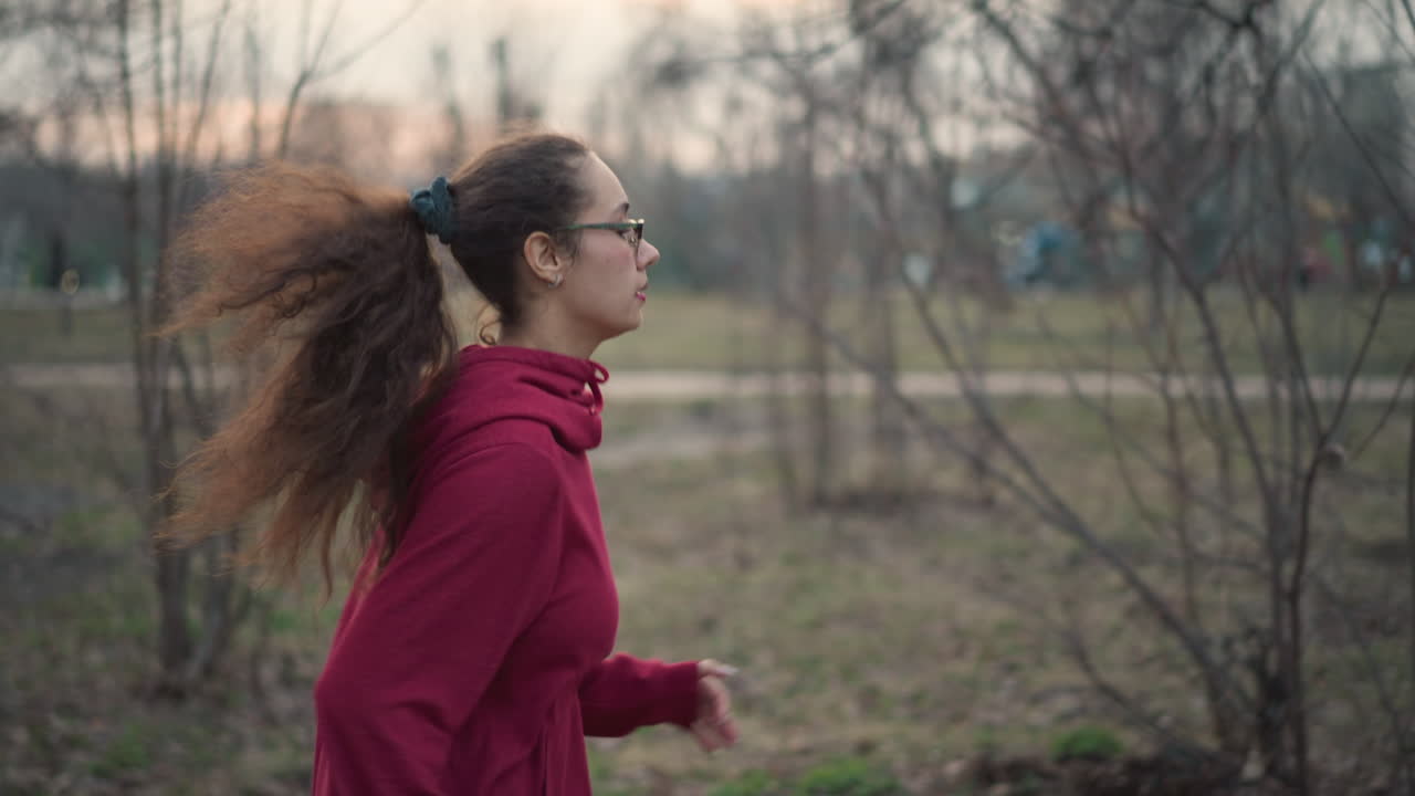 Young Woman Jogging Through Leafless Park Wearing Red Hoodie And Glasses, Ponytail Bouncing, Steady Stride And Focused Expression, Student Jogger On Urban Trail At Dusk, Bare Trees And Muted Sunset
