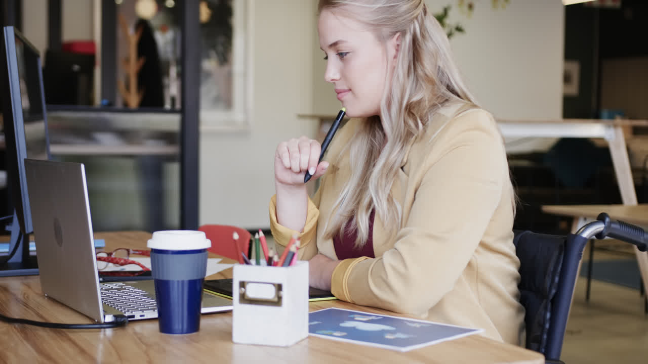 feliz mujer de negocios caucásica casual en silla de ruedas usando computadora portátil en la oficina, en cámara lenta