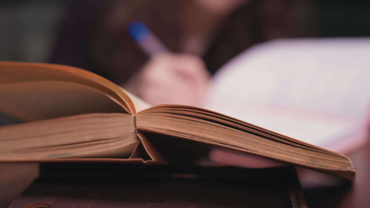 Close up of old brown book open on wooden surface while blurred background shows woman with pen actively writing in notebook, creating cozy and studious indoor atmosphere with warm lighting