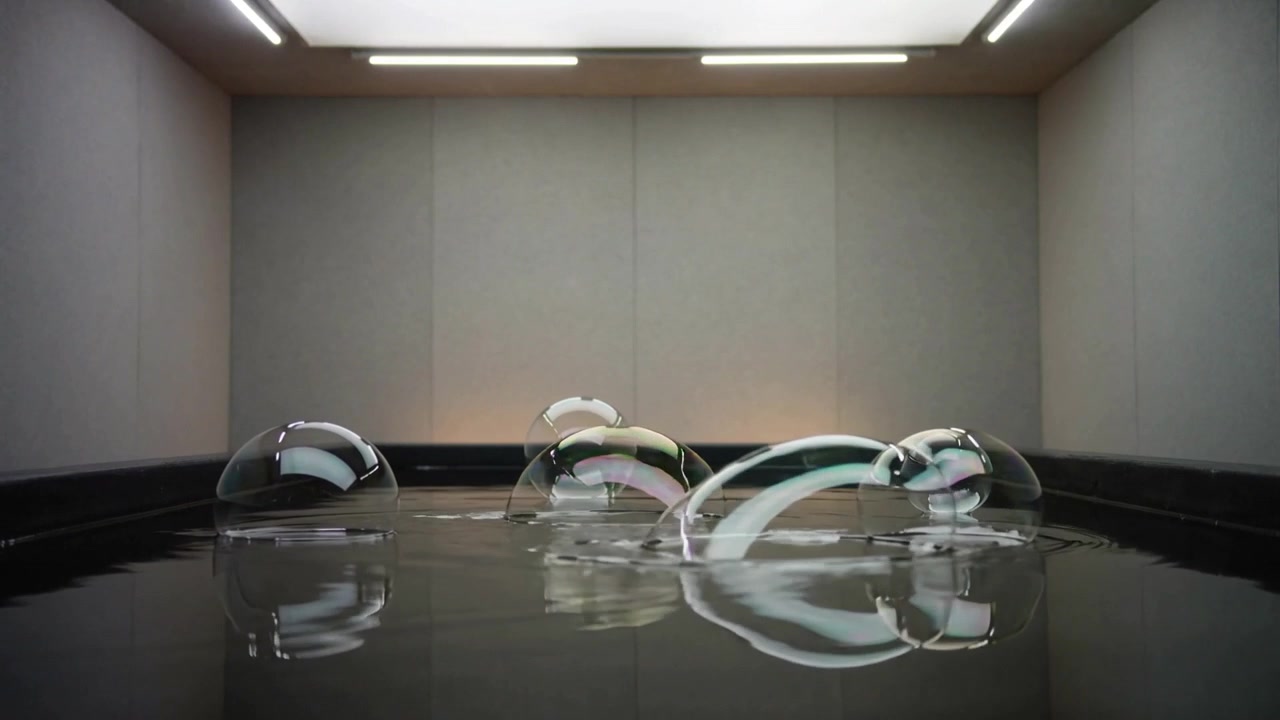 Shifting transparent soap bubbles contacting and merging on shallow basin, reflecting ceiling light