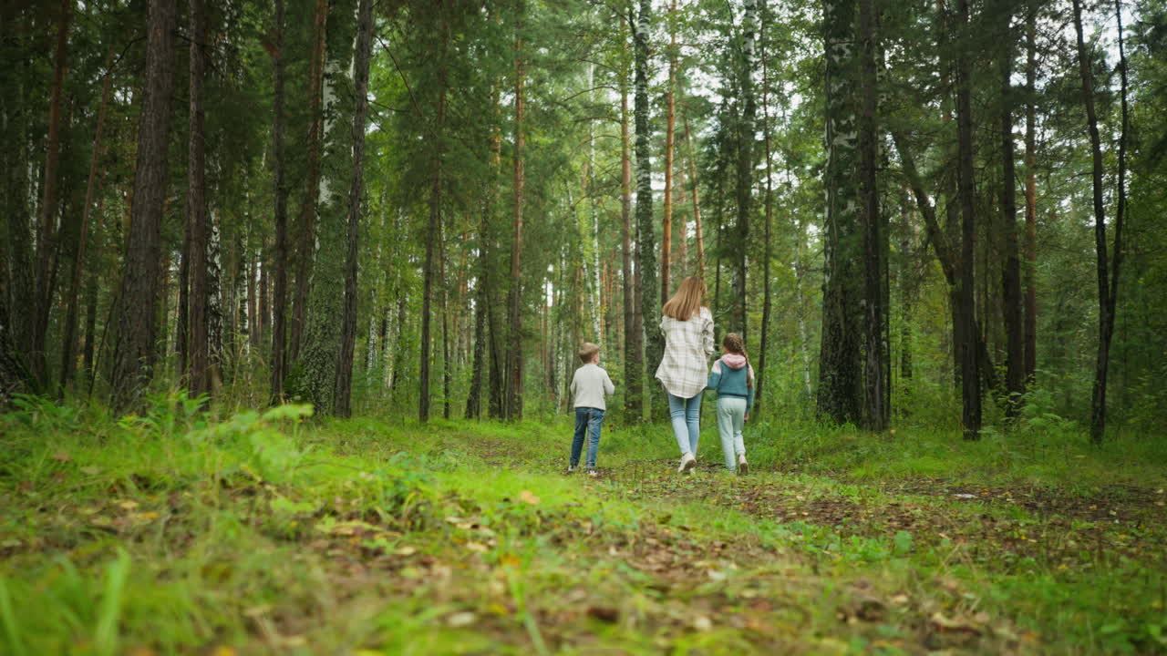 Rear view of mother walking with her two children into dense forest, bonding on nature walk as young boy curiously holds microscope, surrounded by tall trees and fresh greenery