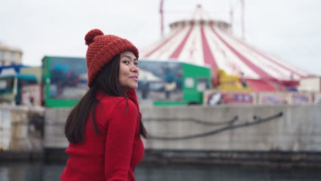 an asian woman smiles at the camera while donning a red beanie, perfectly matching her red turtleneck shirt, in a harbor with a circus-themed backdrop at the port of valencia, spain