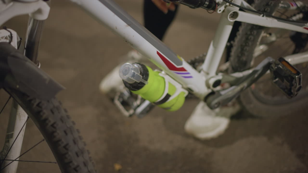 Closeup Of Bicycle Frame And Bottle Reaching Under Frame, Hand Sliding Bright Green Bottle Into Holder, Urban Pavement, Low Light, Soft Bokeh From Streetlights, Subtle Dirt On Tire, Focus On Metal