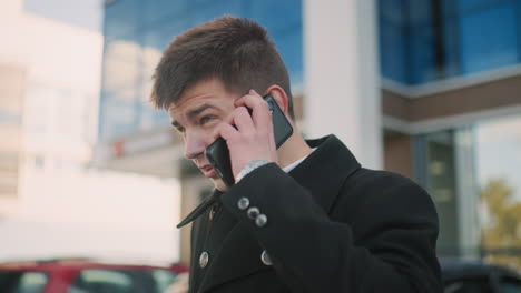 close up of man in black coat talking on two phones interchangeably with focused expression in vibrant urban area featuring modern glass buildings