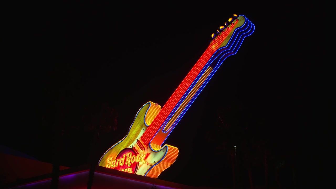 Wide static shot of towering neon-lit electric guitar sign glowing brightly against dark night sky.