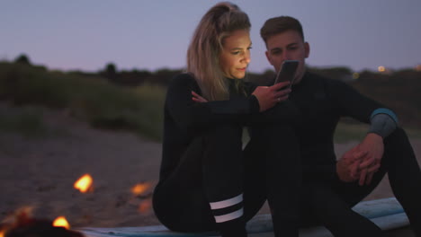 couple sitting on surfboard by camp fire on beach using mobile phone as sun sets behind them