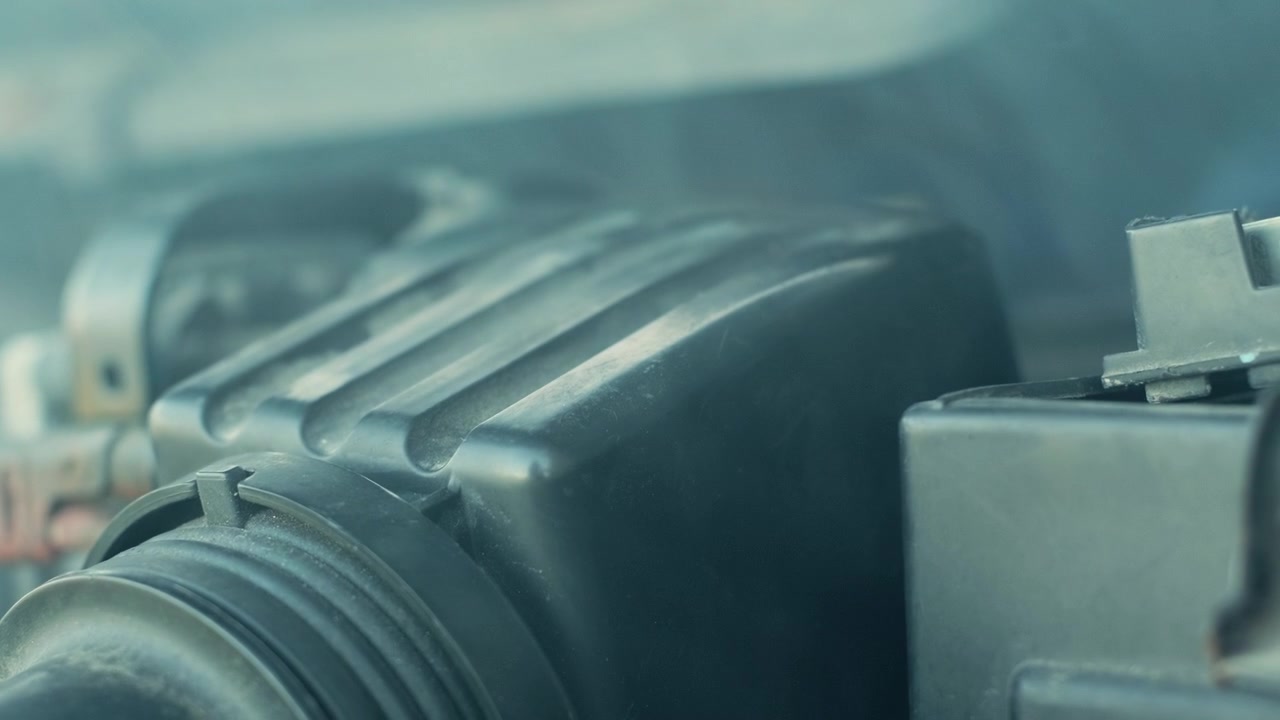 Handheld slow motion shot of engine bay showing smoke drifting around a plastic air intake housing, suggesting overheating or mechanical trouble for auto repair themes.