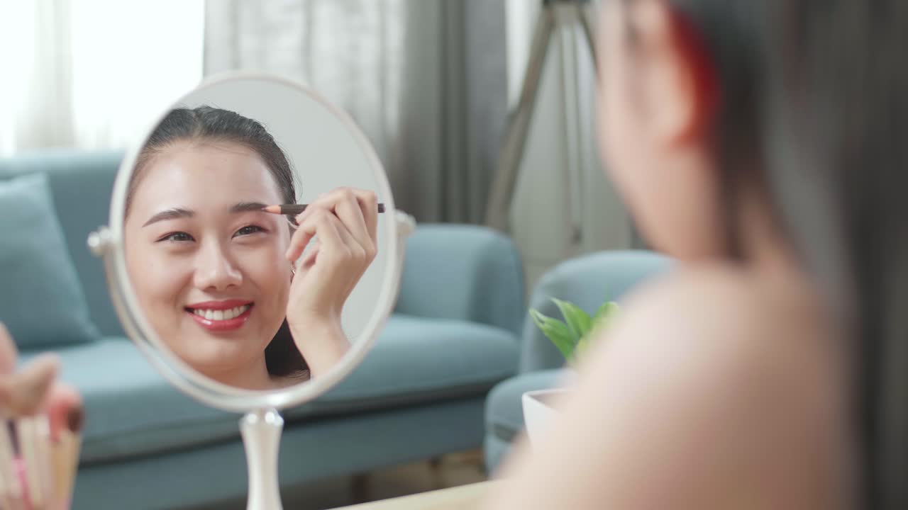 Reflection In A Mirror Beautiful Young Asian Woman Applying Eyebrow Pencil And Smiling While Applying Makeup At Home