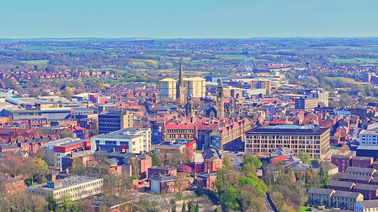 Wide aerial view over Wakefield’s All Saints Cathedral, with traditional brick houses, surrounding greenery, moving traffic, and a clear blue horizon in historic Yorkshire, England.