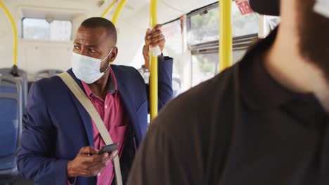 african american man in city wearing face mask, using smartphone in bus