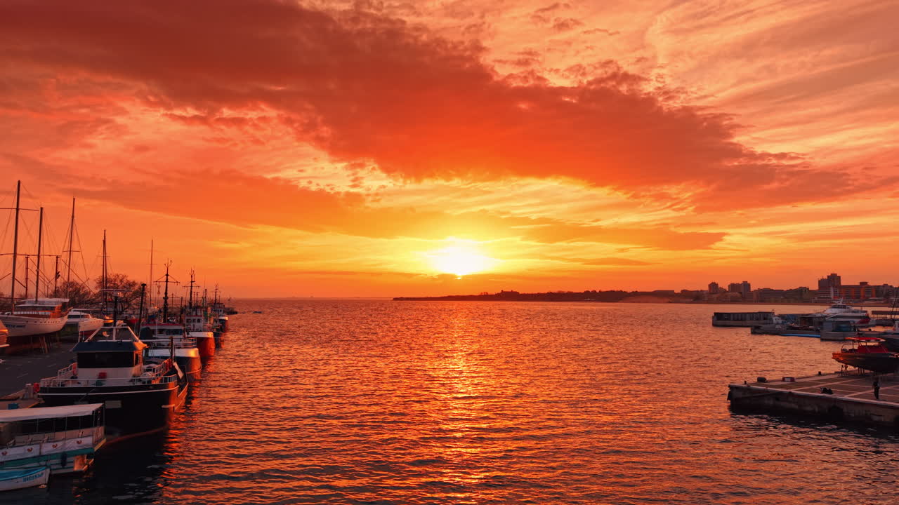 Orange himmel og havlandskab ved solnedgang. Række af både ved kajen. Byens skyline i baggrunden.
