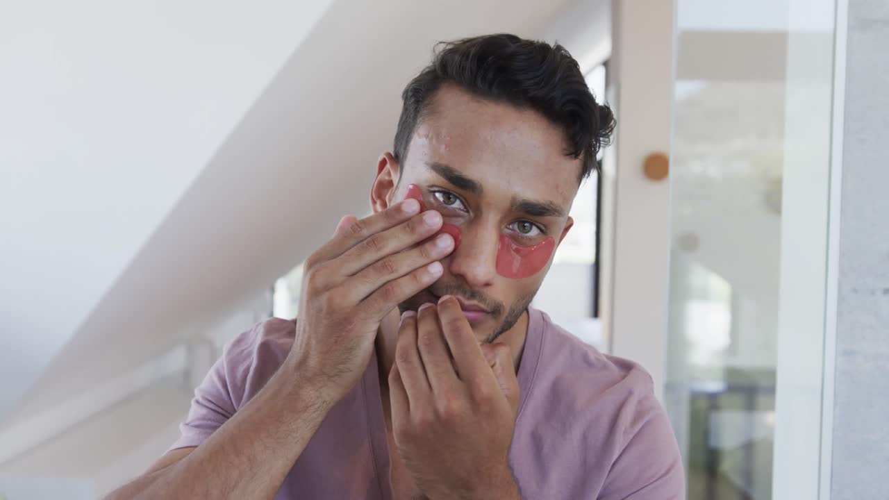 hombre biracial mirándose en el espejo aplicando máscaras debajo de los ojos en el baño, cámara lenta