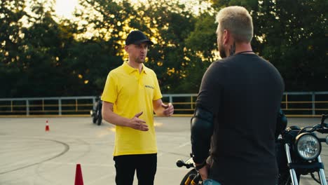 A-male-instructor-in-a-yellow-T-shirt-and-cap-tells-a-biker-with-a-beard-about-the-rules-of-driving-a-motorcycle