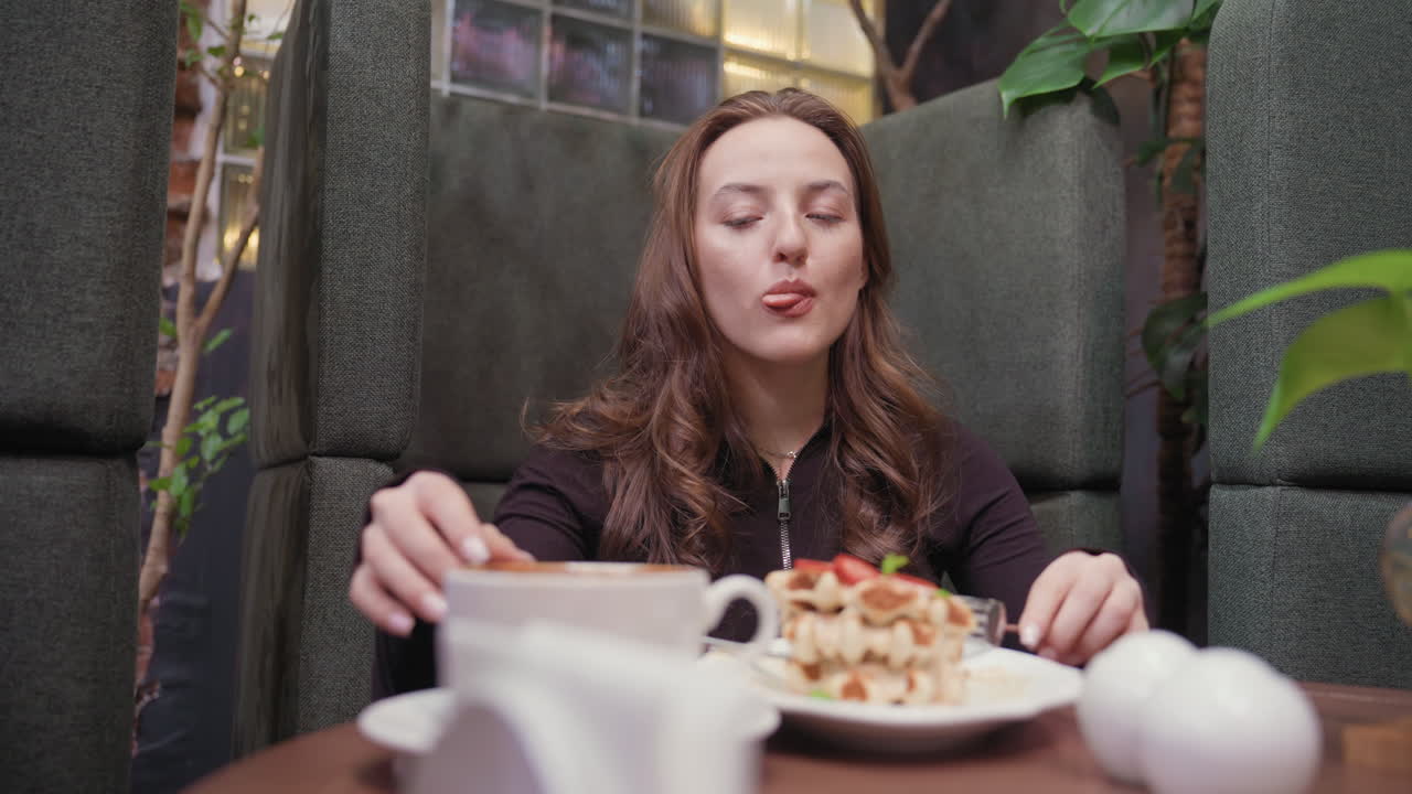 Lady enjoys delicious waffles while leaning back comfortably in cozy booth, sipping latte beside decorative plants and warm interior elements, capturing moment of relaxed indulgence and satisfaction