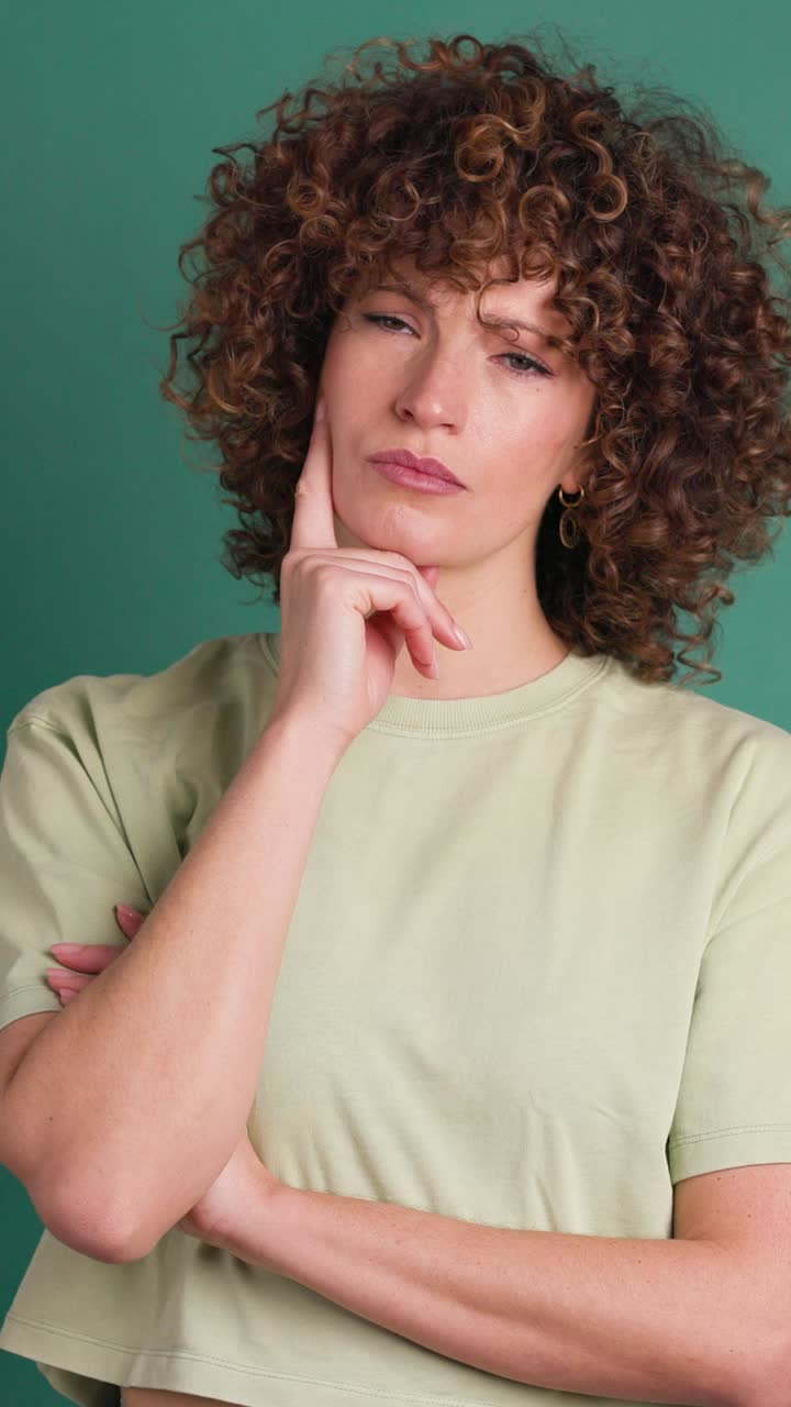 Thoughtful young woman touching face and smiling on green background. Vertical orientation