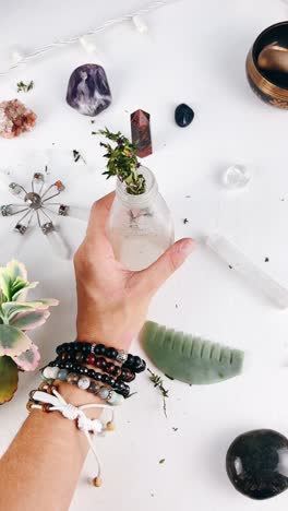 hand holding a small glass bottle with a plant, surrounded by crystals and jewelry on a white surface