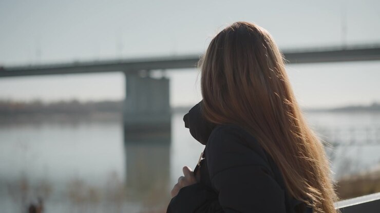Una donna con i capelli lunghi sta in piedi in riva al fiume, indossa una giacca scura e tiene un oggetto vicino al petto. Lo sfondo mostra acqua calma che riflette il cielo e strutture distanti, incluso un ponte.