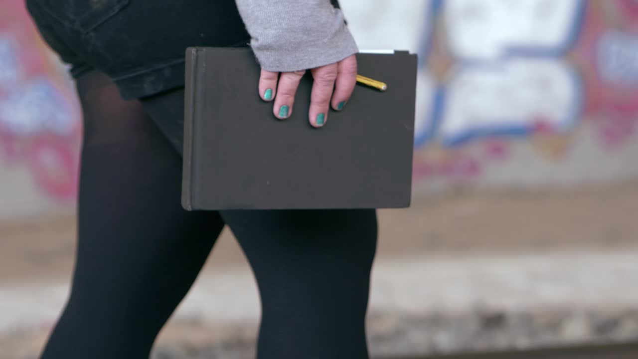 Dívka s modře nalakovanými nehty kráčí se skicákem a tužkou v ruce tunelem plným graffiti.