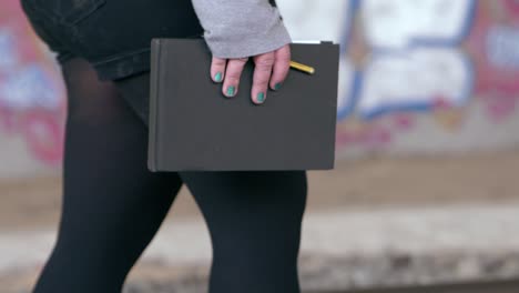 Girl-with-nails-painted-blue-walks-with-her-sketchbook-and-pencil-in-her-hand-through-a-tunnel-full-of-graffiti