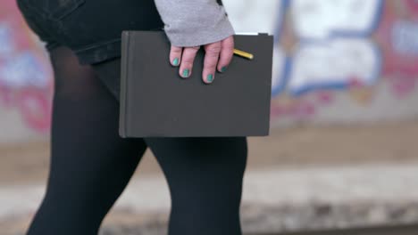 una chica con las uñas pintadas de azul camina con su cuaderno de bocetos y un lápiz en la mano a través de un túnel lleno de graffitis