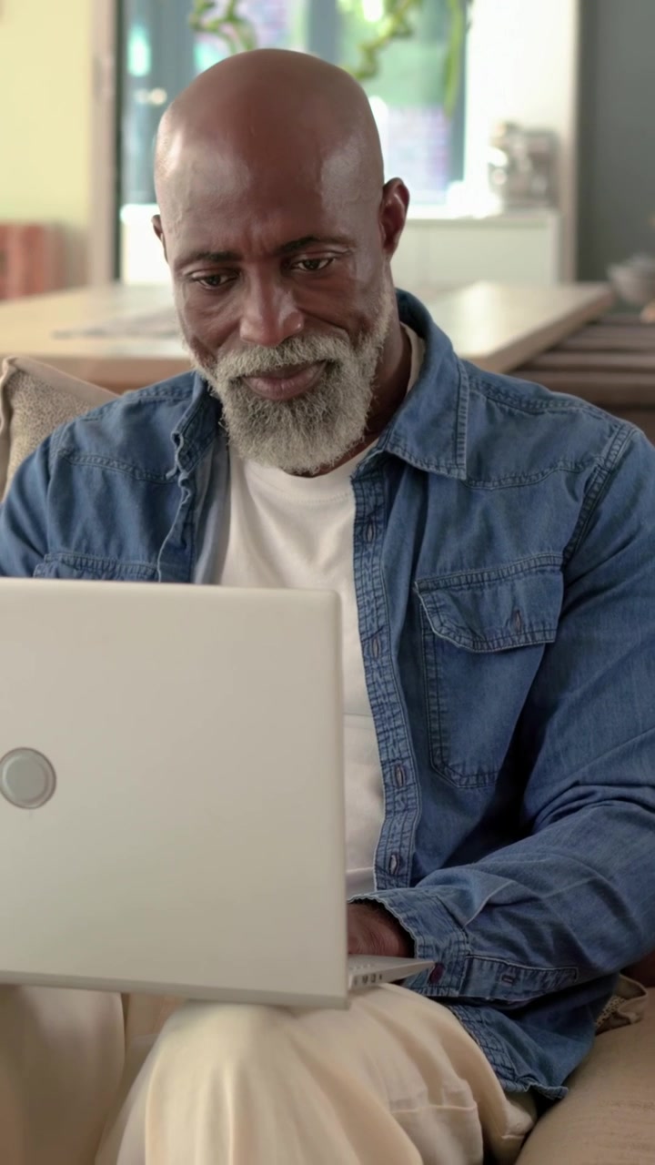 Ældre afroamerikansk mand taster på bærbar computer på sofaen, efter bevægelse ved skydedør, smilende og tastende