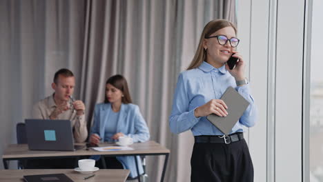 happy self-confident business lady having phone conversation standing near the office window. a pair of colleagues sit at desk behind discussing important job issues.