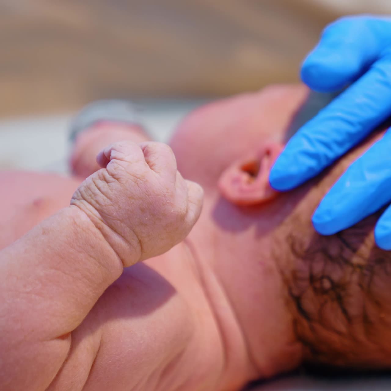 Hand in blue latex glove strokes the head of a newborn. Caucasian baby lies naked on the table. Close up