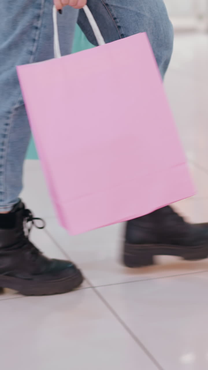 바쁜 쇼핑몰에서 상점을 지나가는 동안 분홍색과 민트 쇼핑 가방을 들고 검은색 부츠와 청바지를 입은 스타일리시한 여성의 아래쪽 다리 시각, 다른 쇼핑객들이 지나가는 장면에 움직임을 추가합니다.