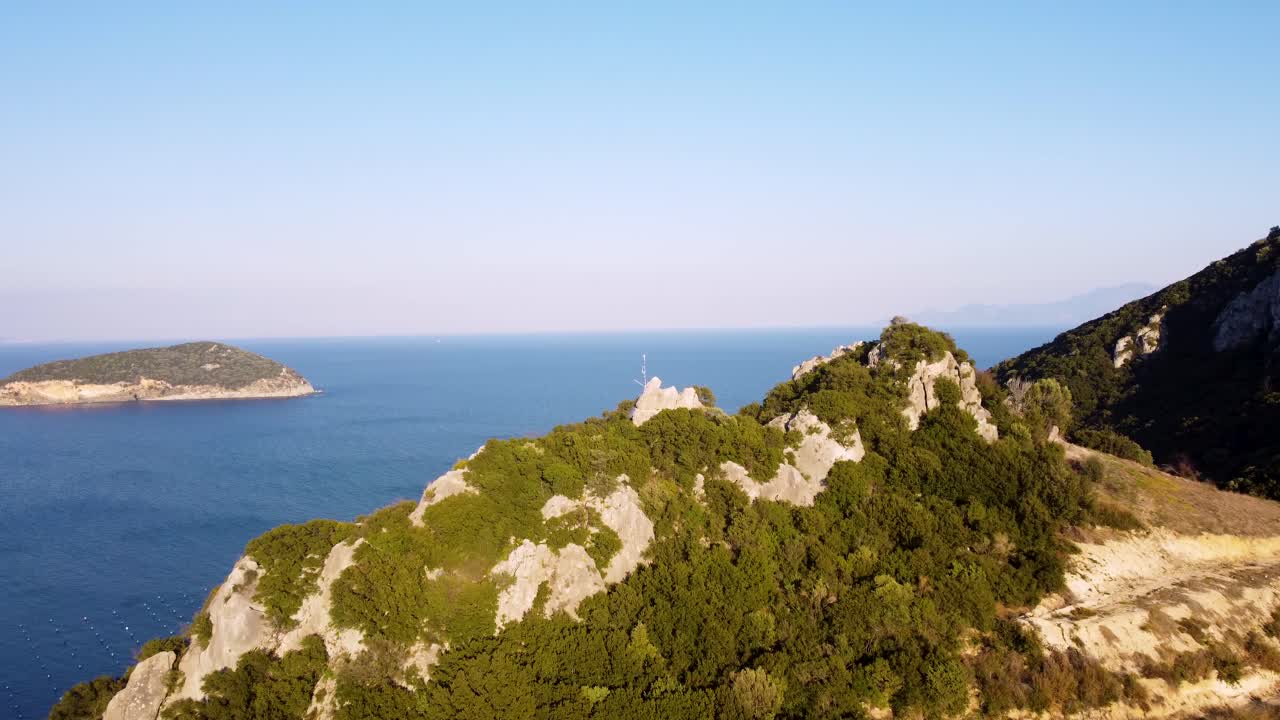 Aerial approaching a wind-torn Greek flag on top of a mountain next to the ocean at sunset.