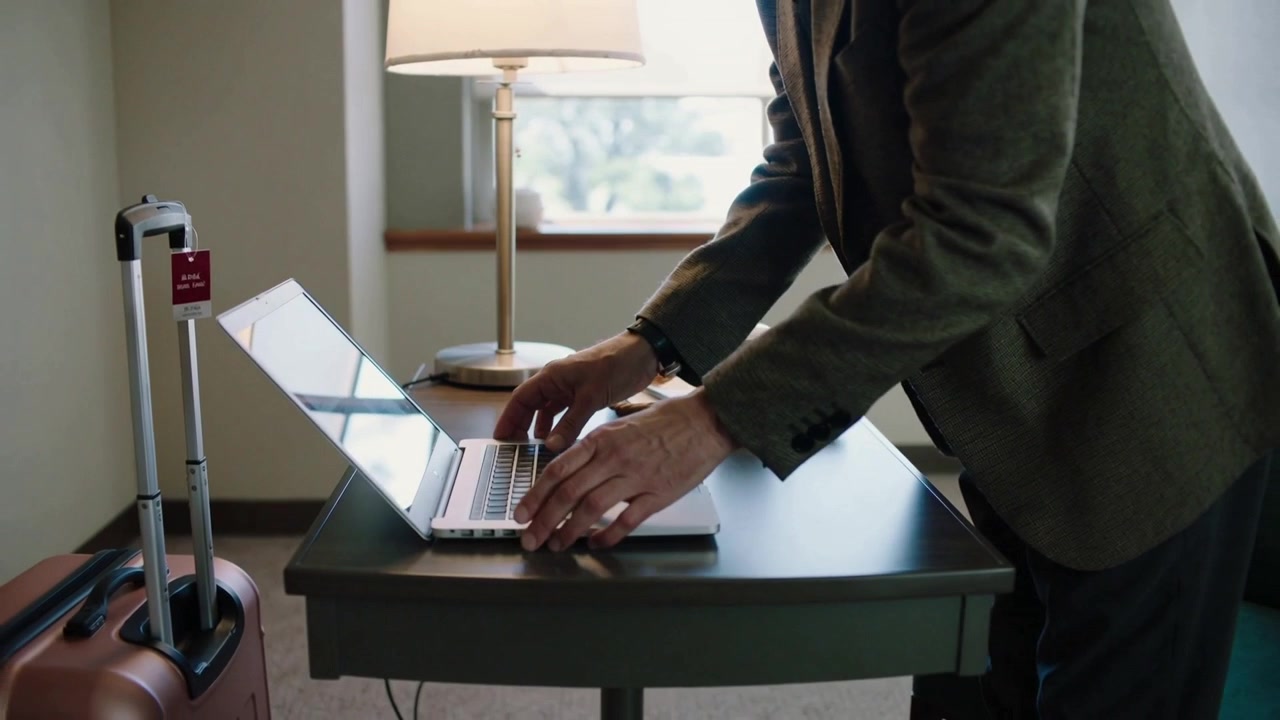 Arriving with red-tagged suitcase, blazer-clad man opening laptop and typing for work at hotel desk